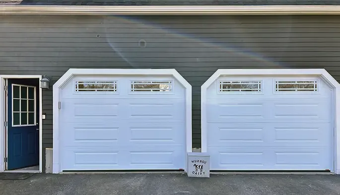 Two white garage doors with small windows on a gray house, adjacent to a blue door.